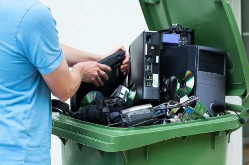 A person wearing a light blue T-shirt is shown loading discarded electronic waste into a large green wheelie bin in an outdoor setting, possibly a driveway or garden area. The waste includes a variety of old electronic items such as a black desktop computer tower, a flat-screen monitor resting on its side, a DSLR camera with attached lens, a small portable radio, and other electronic components and accessories with visible cables and circuit boards. The electronic waste appears used and damaged, with some components partially disassembled. The green bin's lid is open, revealing the mixture of electronic debris inside, set against a background of a paved surface and greenery, indicative of a residential environment. The scene relates to rubbish removal services for electronic or garden waste, consistent with Garden Clearance Beckenham's scope of work in the local area around Beckenham.