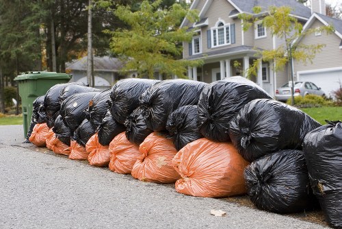 A row of large black and orange rubbish bags filled with waste material, lined up along the edge of a residential street in Beckenham. The bags are stacked on the pavement, close to the curb, with the black bags placed on top of the orange ones. Behind the bags, there is a green wheelie bin, and further in the background, there are trees with green leaves and a row of detached houses featuring light-colored facades, gabled roofs, and bay windows. The scene is outdoors during daylight hours, with natural light illuminating the area. The image visually depicts typical rubbish collection preparation, which Garden Clearance Beckenham might assist with as part of their garden clearance services, emphasizing their capacity for managing various types of waste disposal in the local area.